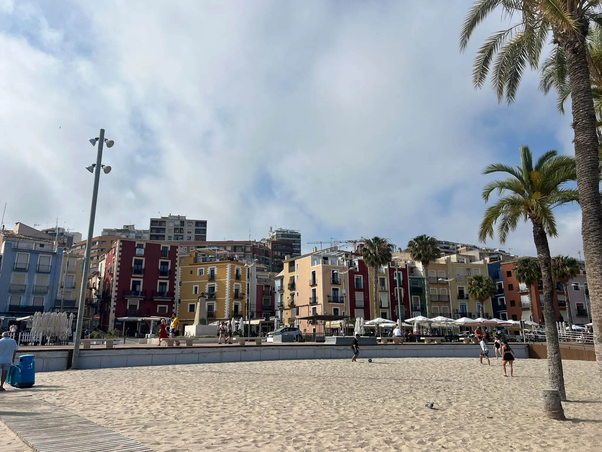 Sunny daylight view of Playa Centro in Villajoyosa — people near wooden walkway, backdrop of beach promenade with outdoor restaurant terraces and rows of iconic colourful historic houses