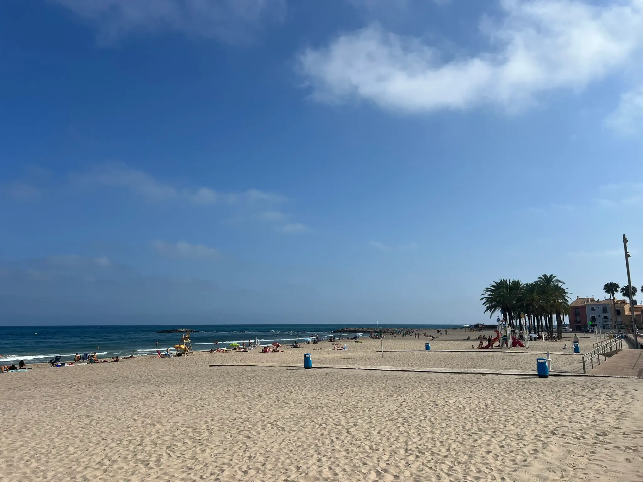 Low-angle perspective of wide sandy Playa Centro in Villajoyosa — wooden pedestrian walkways on sand, long promenade with tall palm trees and traditional colourful houses under bright sky