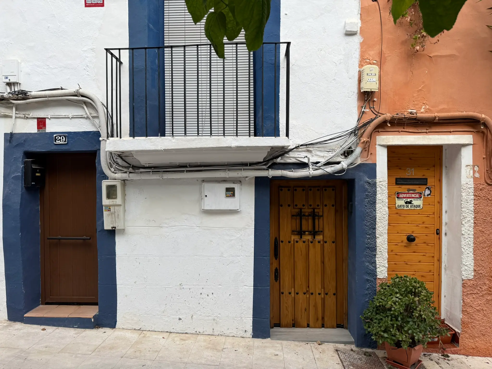 Ground floor facades of houses 29 and 31 in Villajoyosa — white wall with dark blue trim, wooden doors, orange building, potted plant, funny warning sign on yellow door